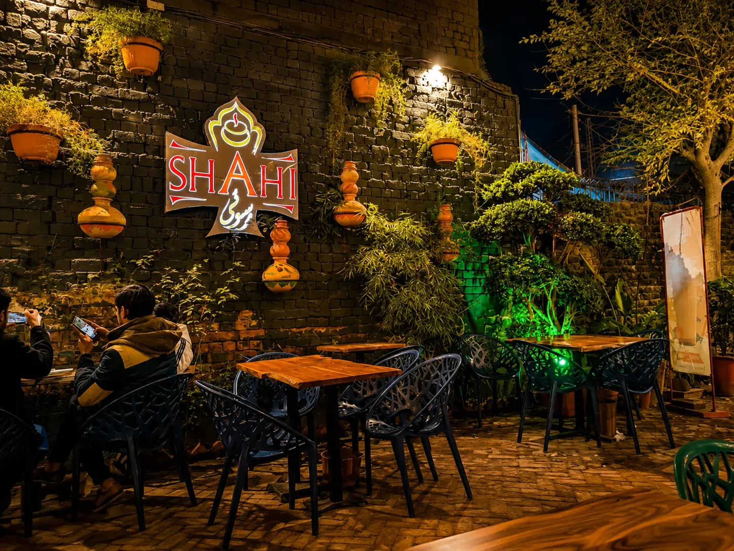 Guests at the outdoor tables at night, the lit Shahi Rasoi sign on the stone wall behind